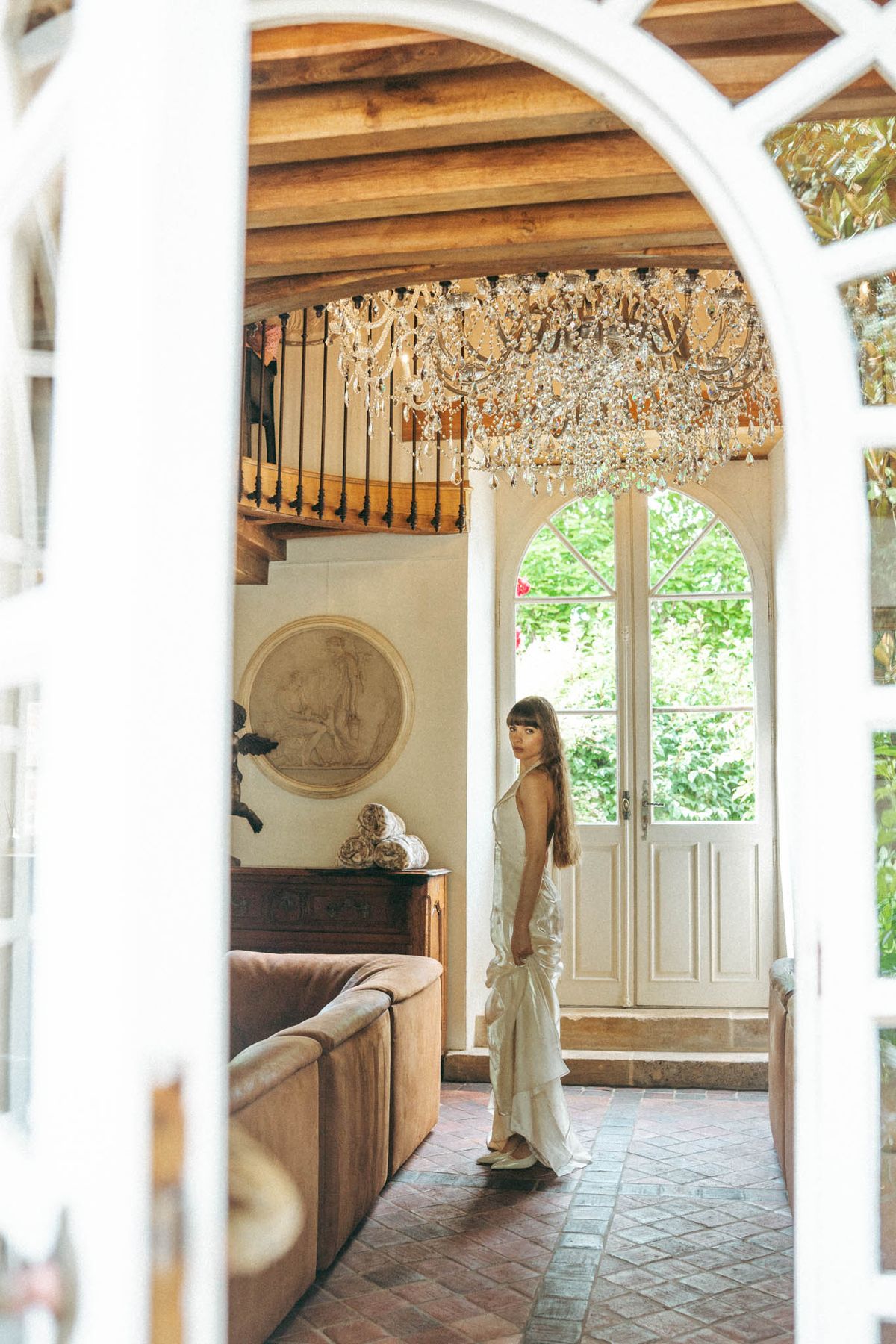 Bride in ivory slip gown framed by arched doorway in rustic interior with chandelier beams and boiserie