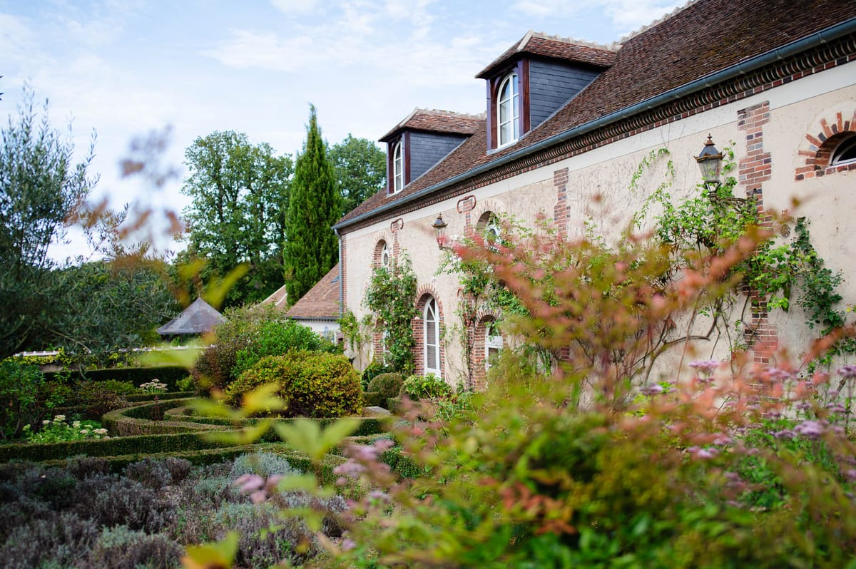 French country estate with red brick arches, parterre garden with mauve perennials, and climbing plants