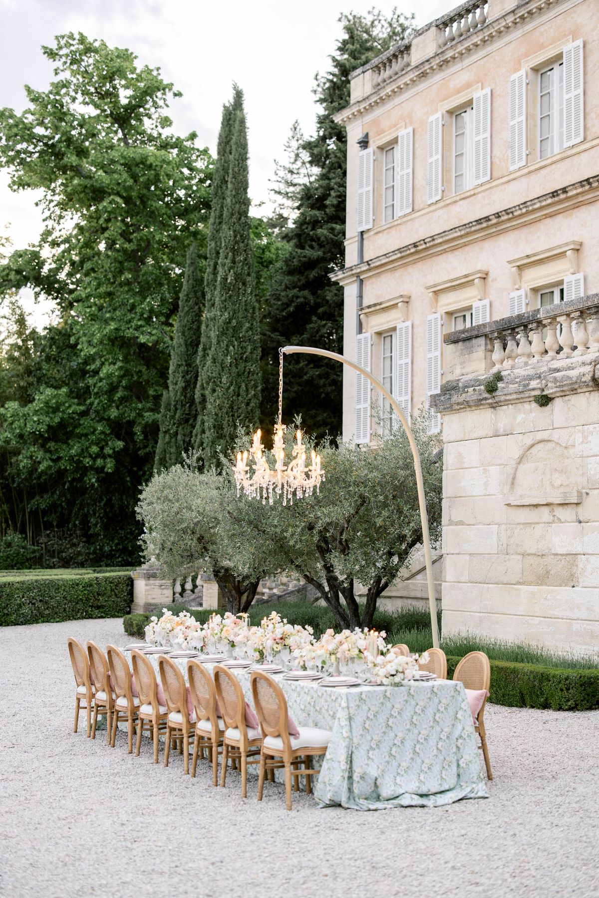 Long outdoor reception table with mint green linen and blush florals beside chateau, gold chandelier overhead