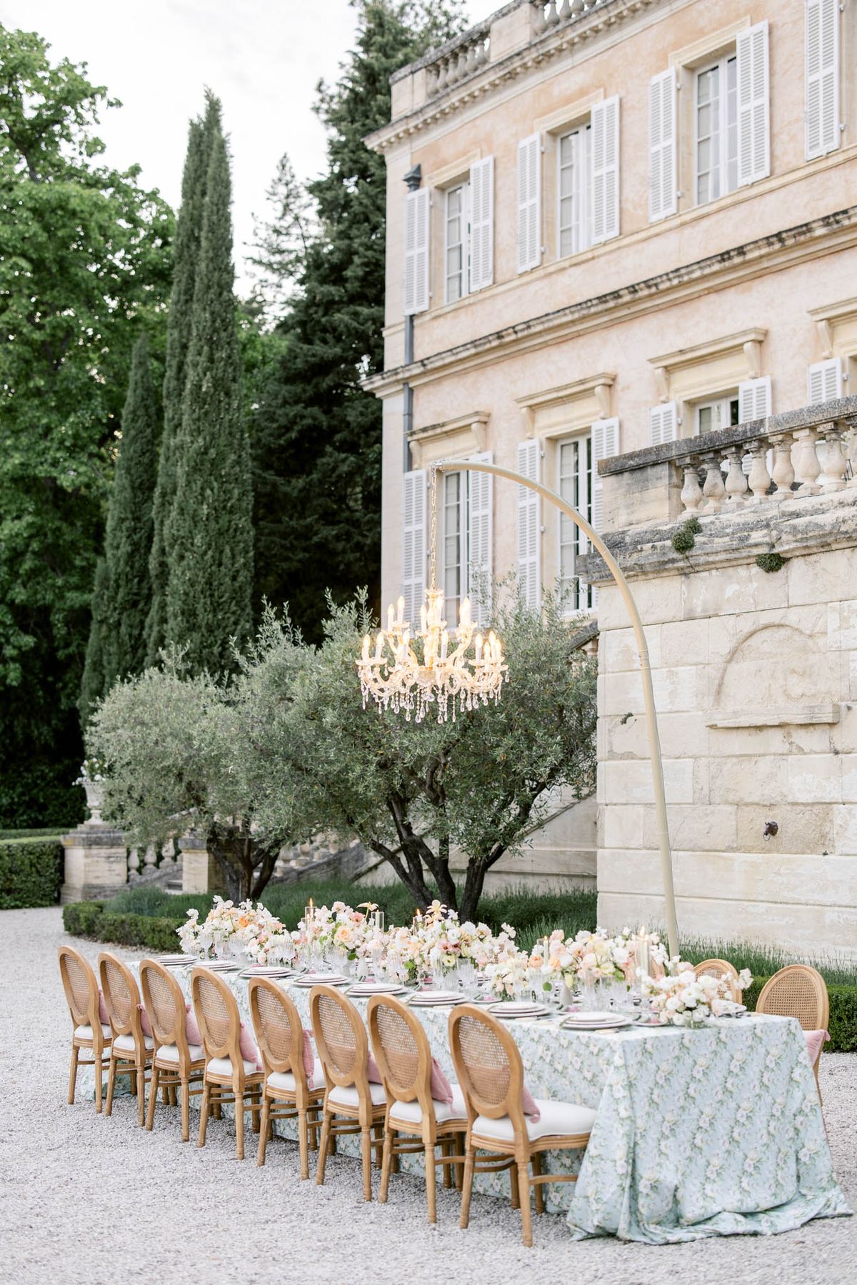 Long reception table with sage green linen and blush floral arrangements before French chateau facade with crystal chandelier