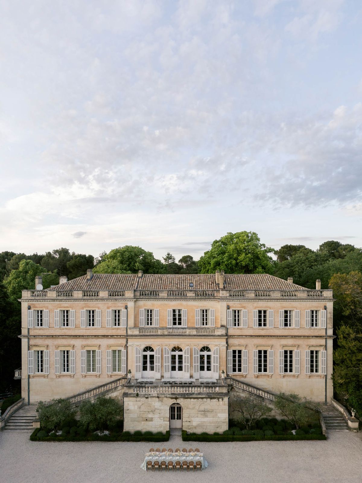Aerial honey-stone chateau with terracotta roof and long reception table set on gravel forecourt before balustraded stairs