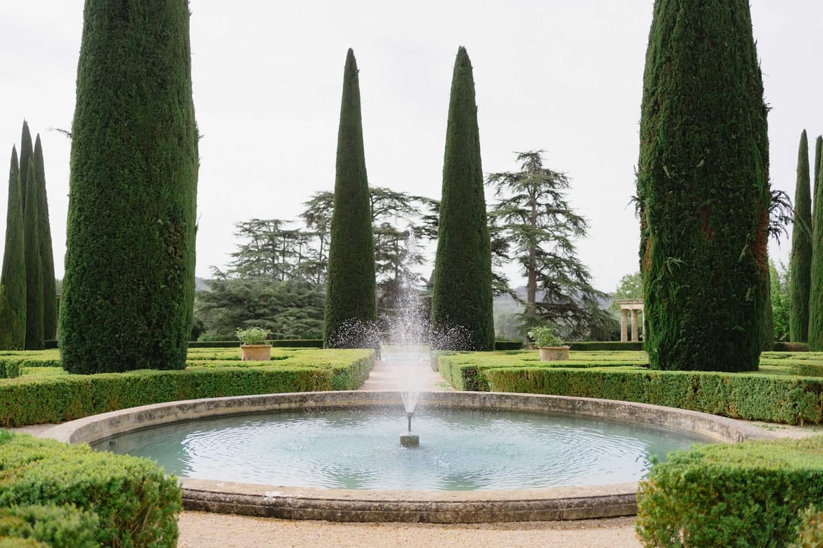 Formal French parterre garden with stone fountain, boxwood hedges, cypress trees, and colonnade