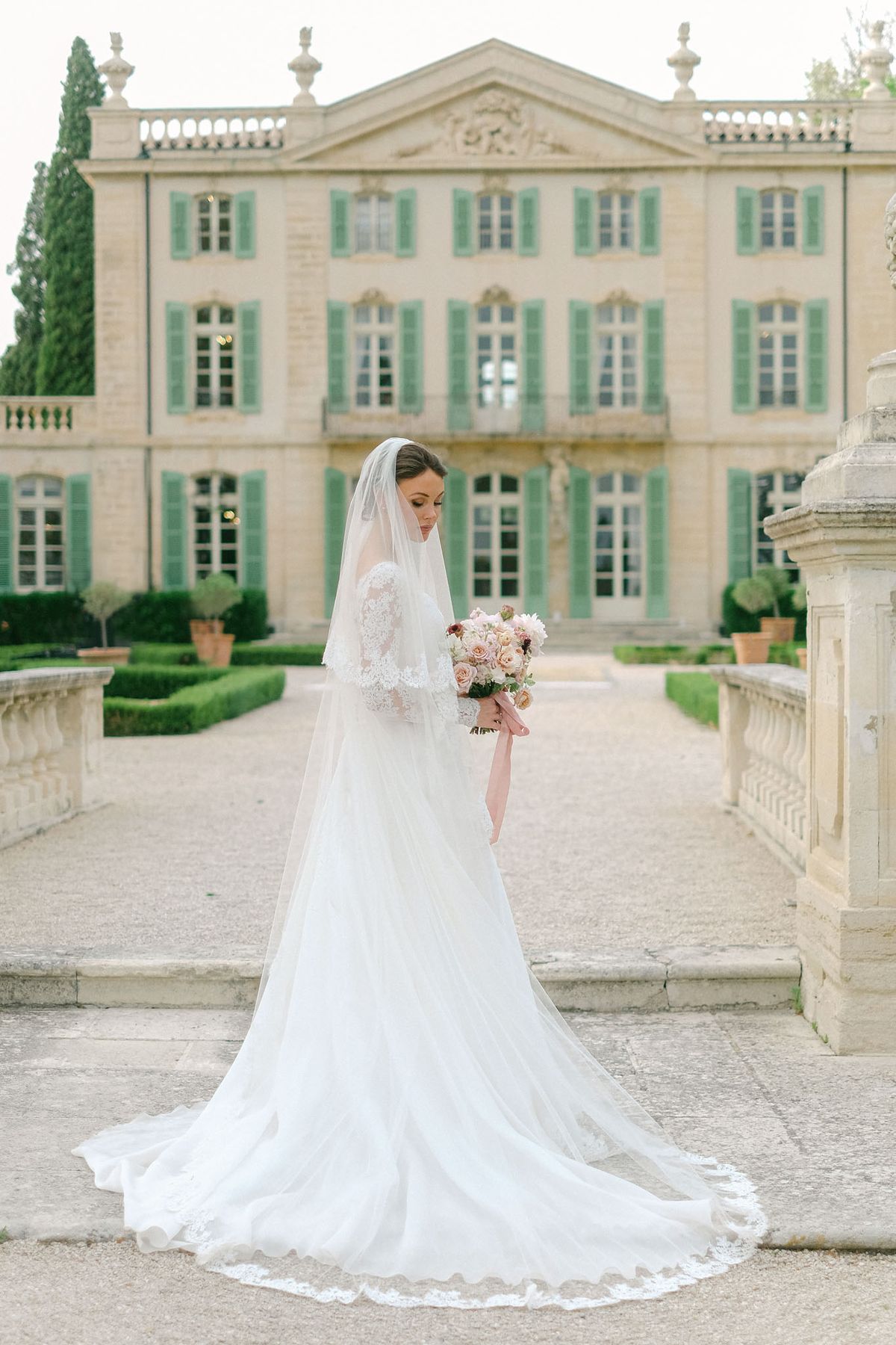 Bride in lace-sleeve ballgown with dusty rose bouquet on chateau terrace with sage shutters and topiary