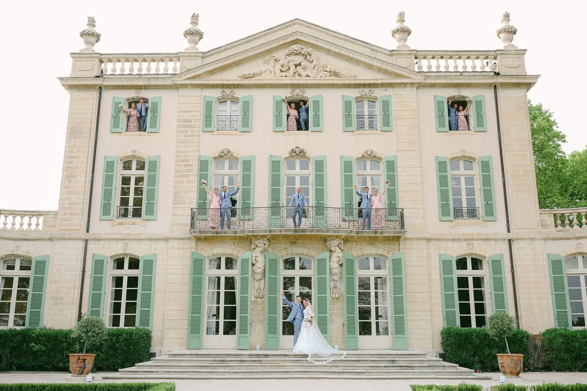 Bridal party posed across three levels of pink chateau facade with bride and groom on steps and attendants at windows