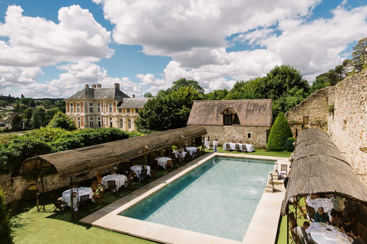 Elevated view of pool courtyard with thatched pergolas, round tables, and classical chateau in background