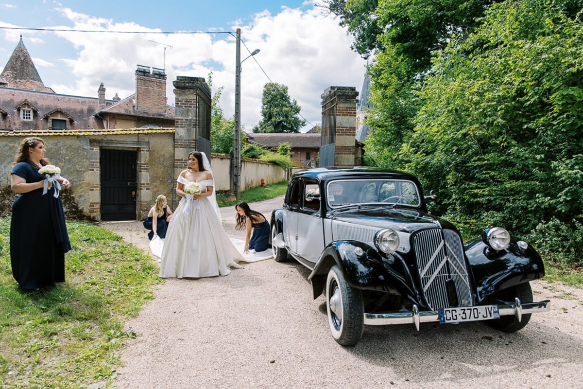 Navy-gowned bridesmaids arranging bride's train beside vintage black Citroen at chateau gates