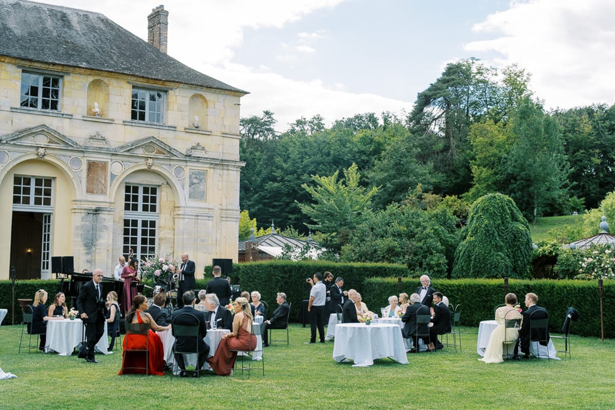 Outdoor wedding reception on a chateau lawn with round white-clothed tables, formally dressed guests, and a grand piano