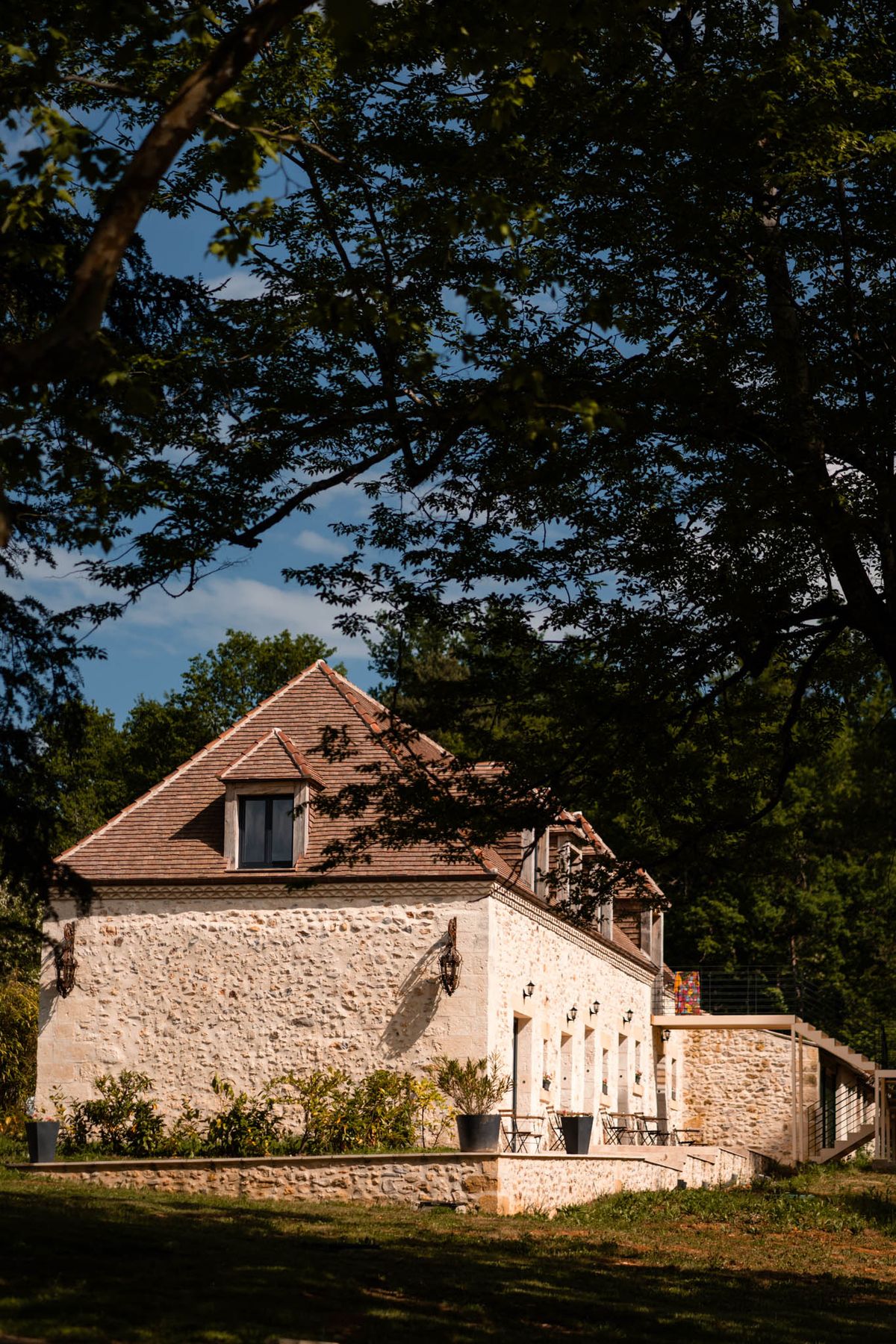 Stone building with terracotta roof and dormer window beneath mature trees with bistro tables on terrace
