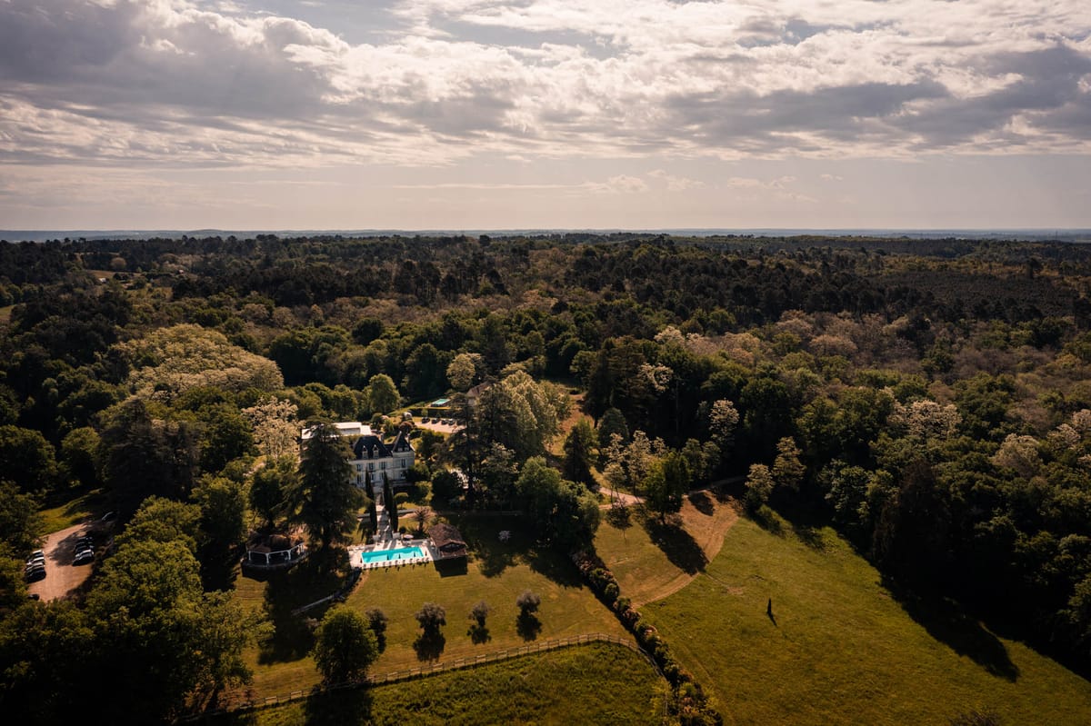 Aerial view of white chateau with mansard roof, turquoise pool, gazebo, and forested estate grounds