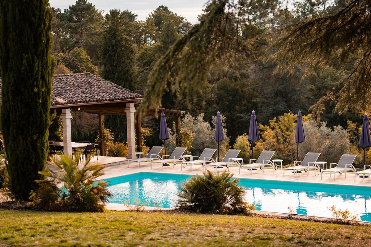 Rectangular pool with grey loungers and navy parasols beside stone-pillared pergola at country estate