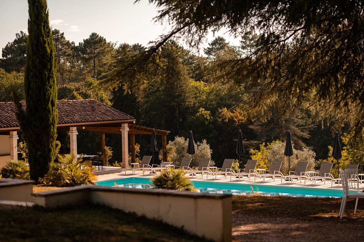 Turquoise pool with loungers and charcoal umbrellas beside stone-columned pergola at golden hour