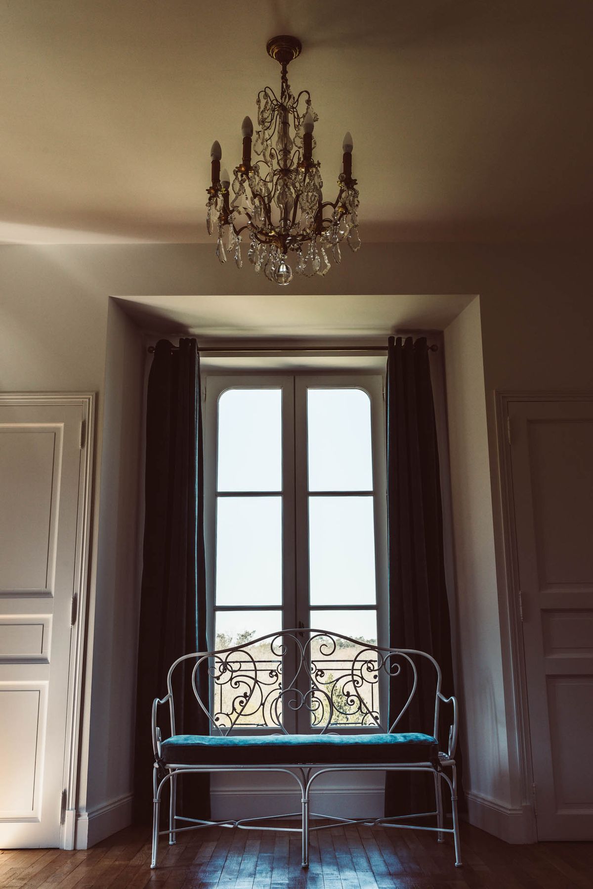 Symmetrical chateau room with wrought-iron bench, teal cushion, arched French doors, and brass chandelier