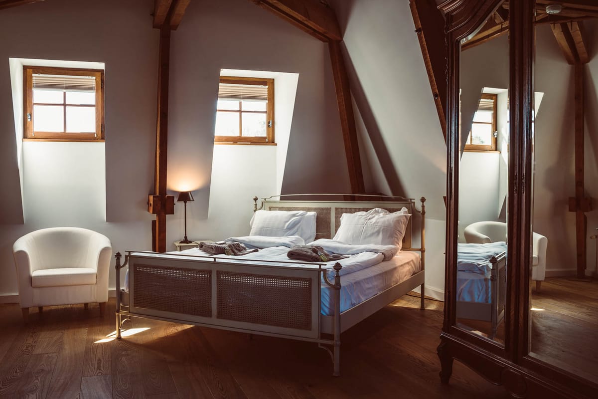 Attic guest bedroom suite in a chateau with exposed wood beams, cane brass bed, white linens, and dormer windows