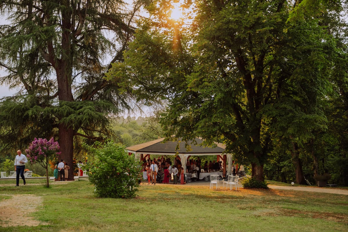 Guests mingling under wooden pavilion with golden afternoon light and lilac bush in bloom
