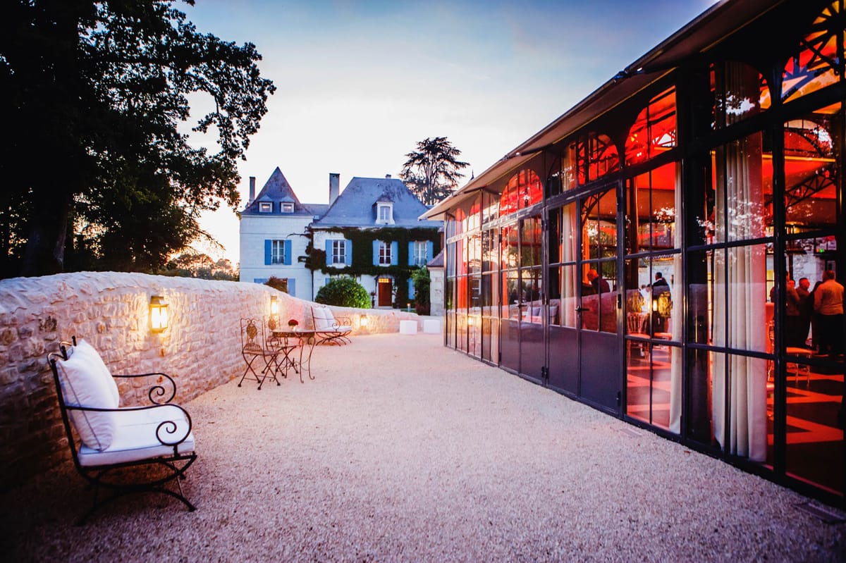 Chateau with blue shutters and glass orangery pavilion glowing red at dusk beside wrought-iron seating
