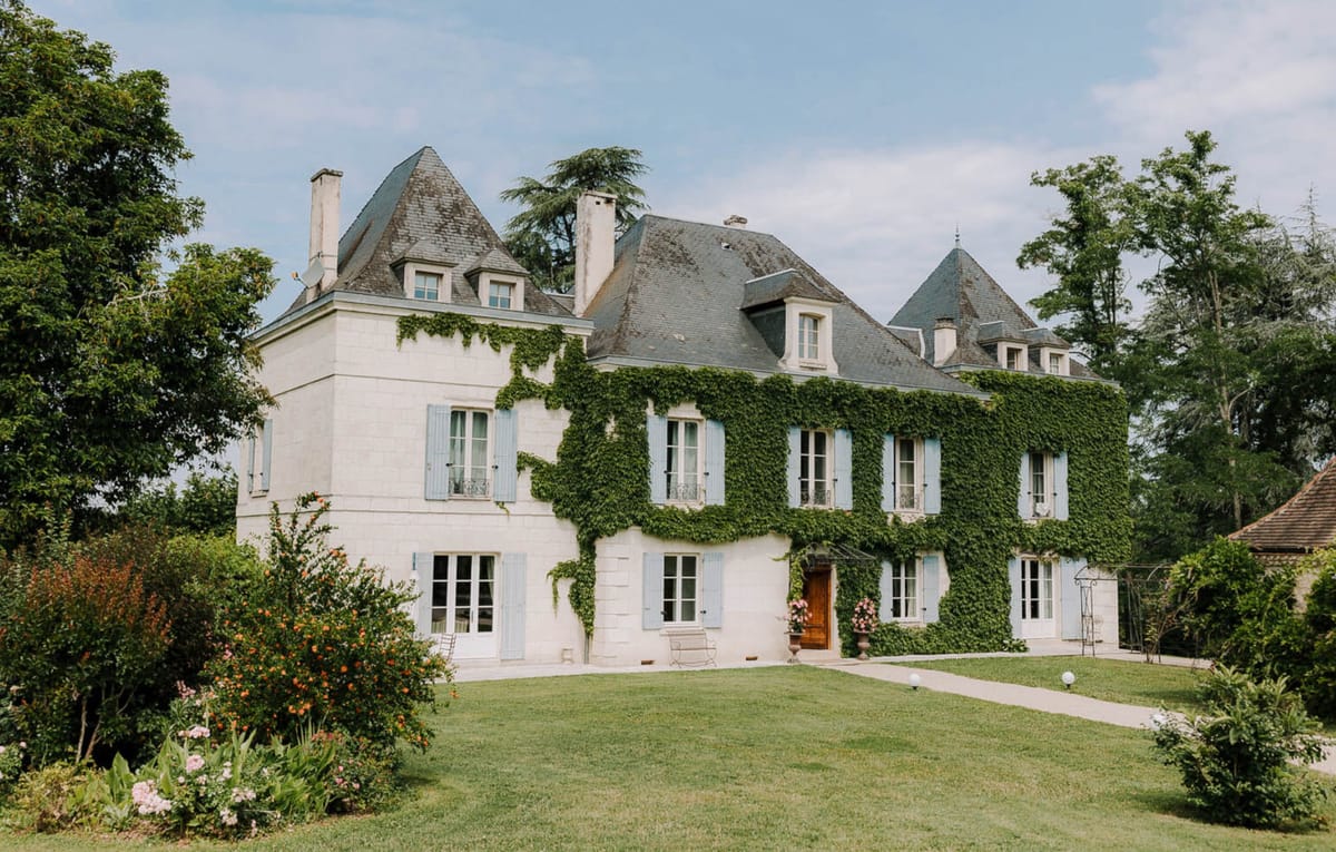 Cream stone chateau with slate turrets, blue shutters, ivy facade, and flowering garden beds on lawn