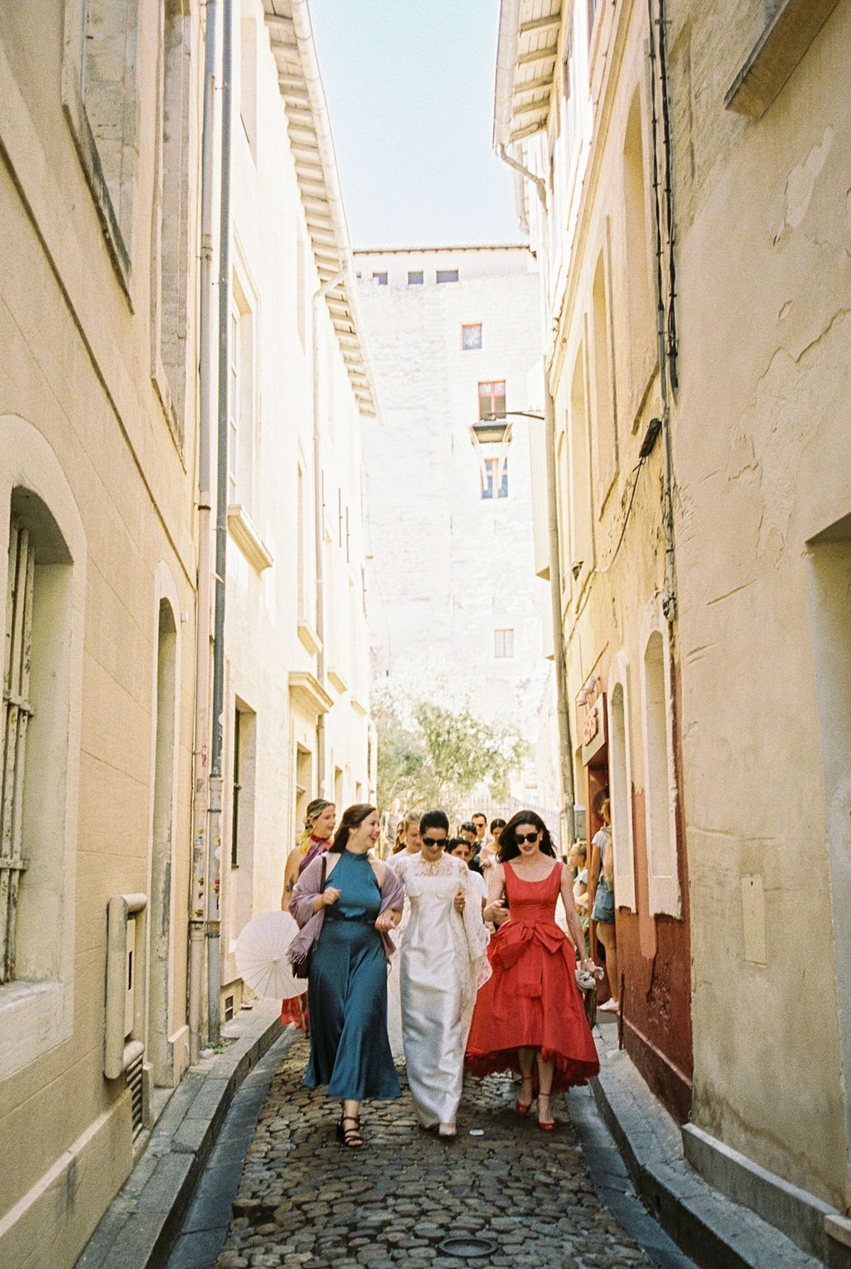 Bride walking cobblestoned Provencal lane with bridesmaids in teal and red gowns