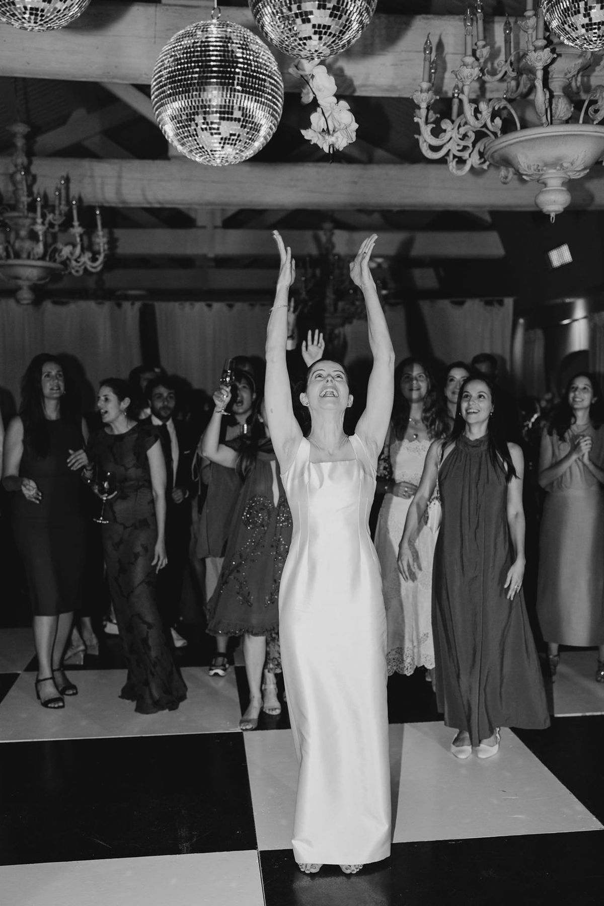 Bride dancing arms raised under disco balls and chandeliers on checkered floor