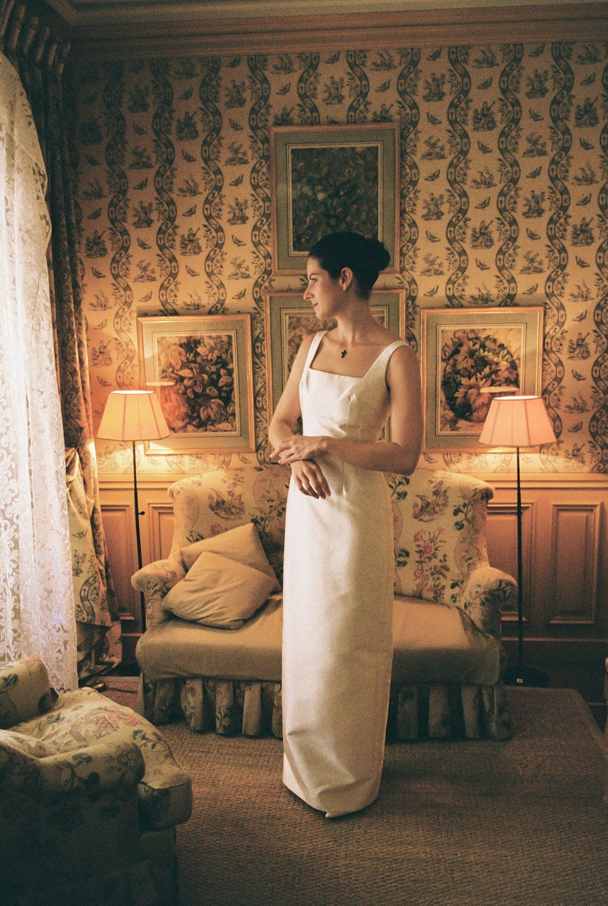 Bride in silk dress fastening pendant in toile-papered chateau bedroom