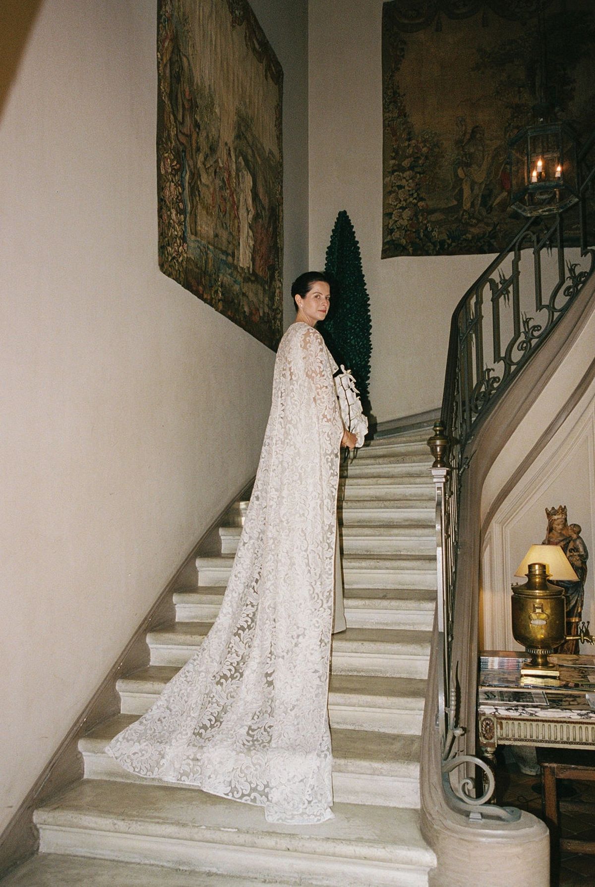 Bride on stone staircase with lace cape trailing past tapestry and iron banister