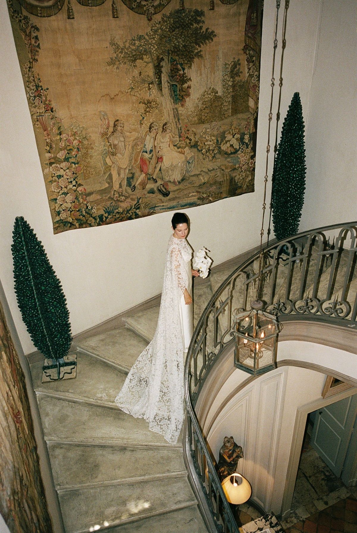Bride descending stone spiral staircase with orchid bouquet beside antique tapestry