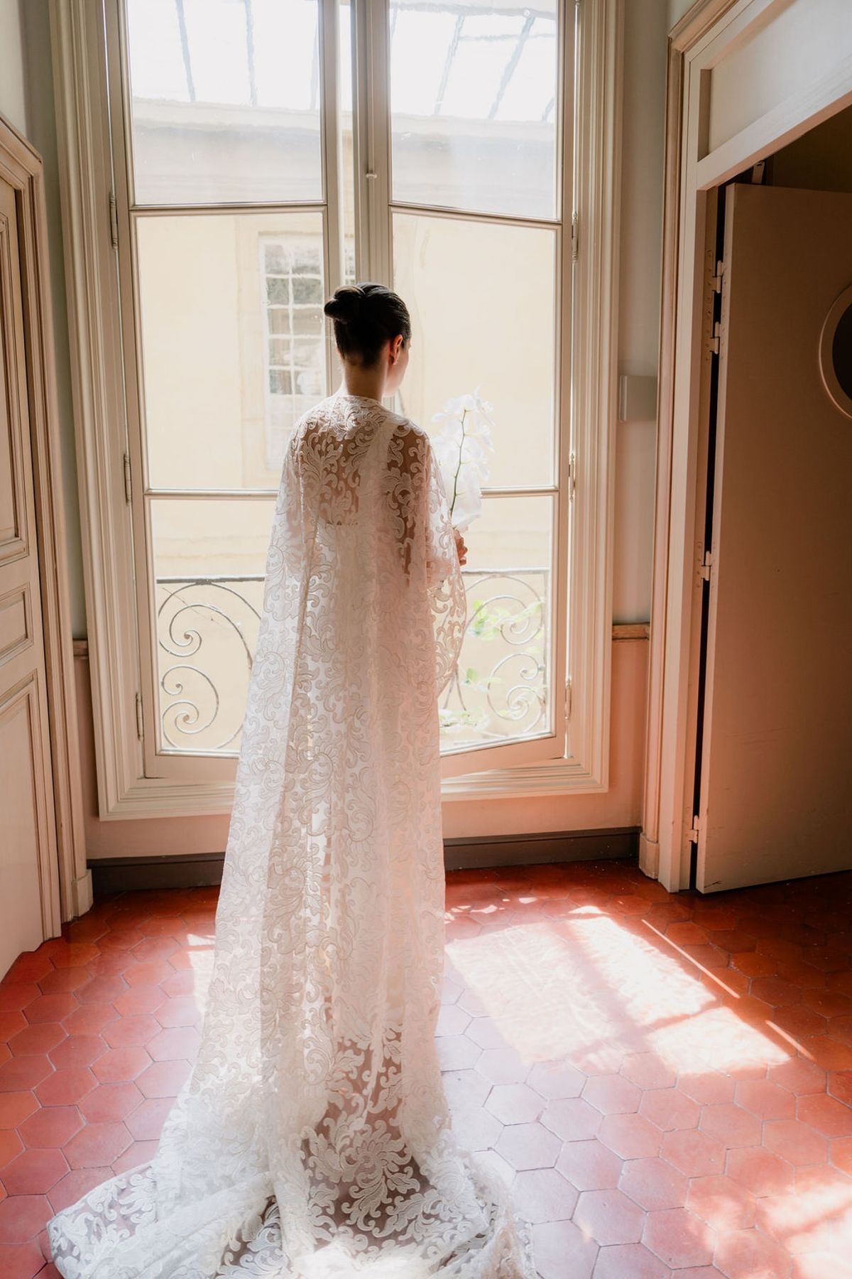Bride at French doors holding orchid stem with lace cape on terracotta tiles