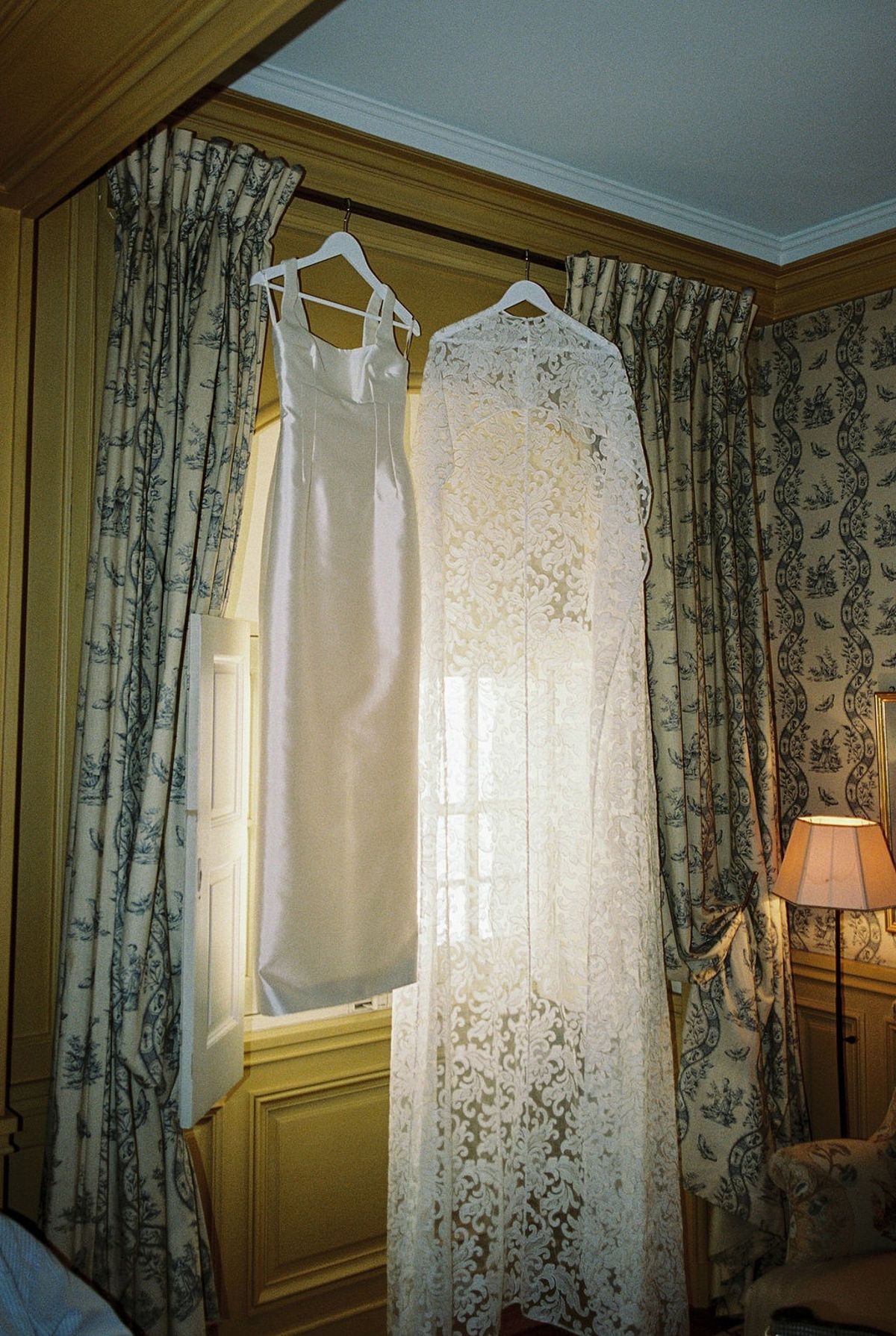 Wedding dress and lace cape hanging at window between toile curtains