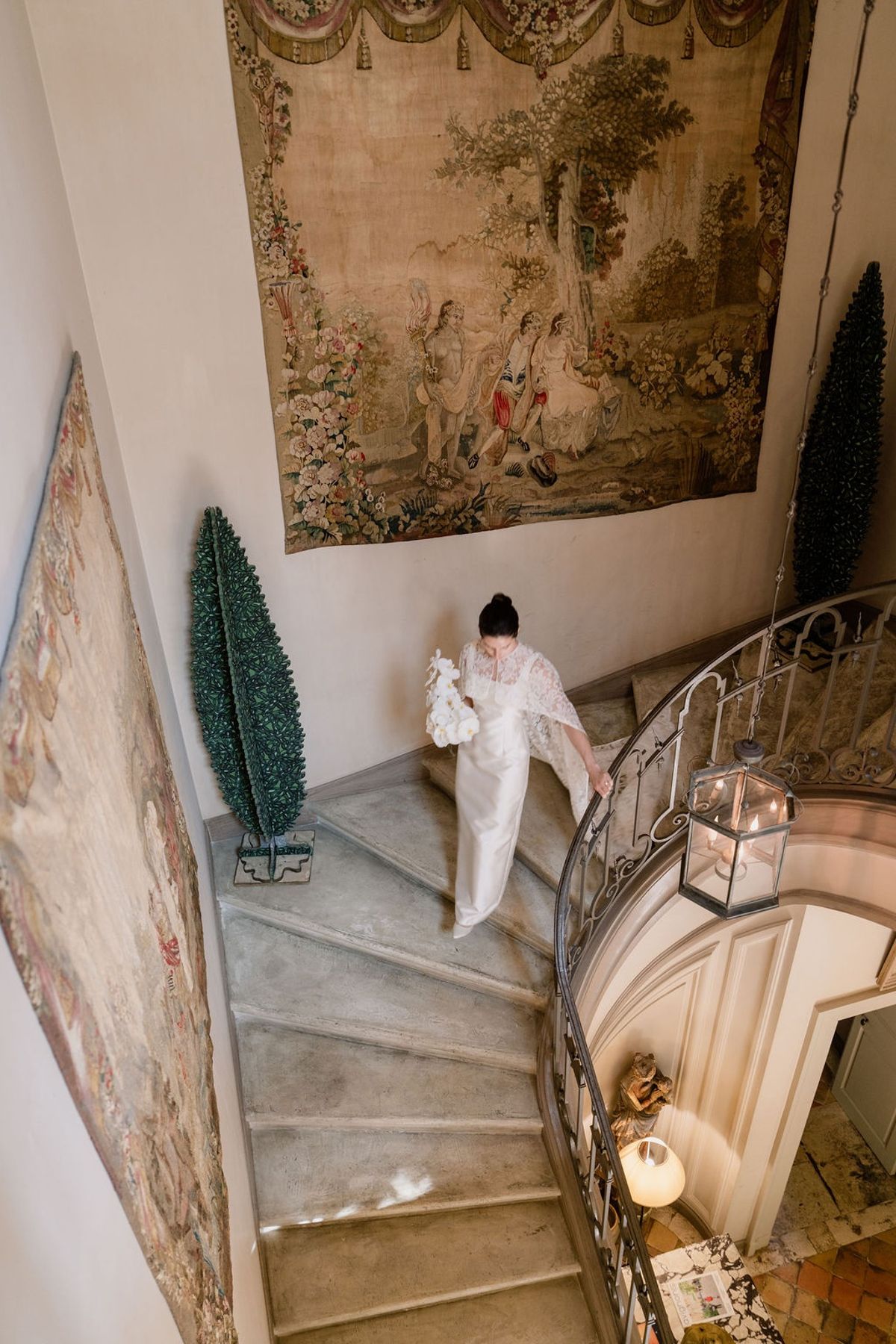 Bride walking down curving stone staircase with orchid bouquet and lace cape