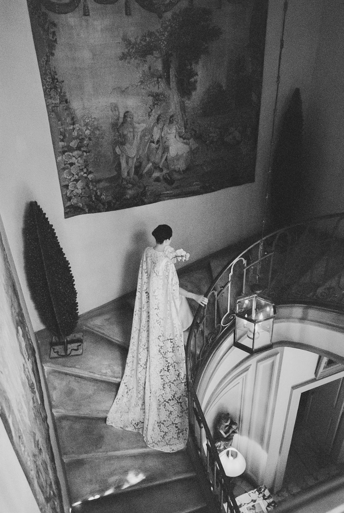 Bride from above ascending stone staircase with lace cape under tapestry