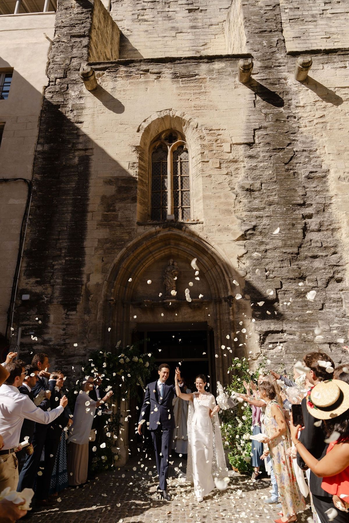 Newlyweds exit medieval church under petal toss from guests in sunlight