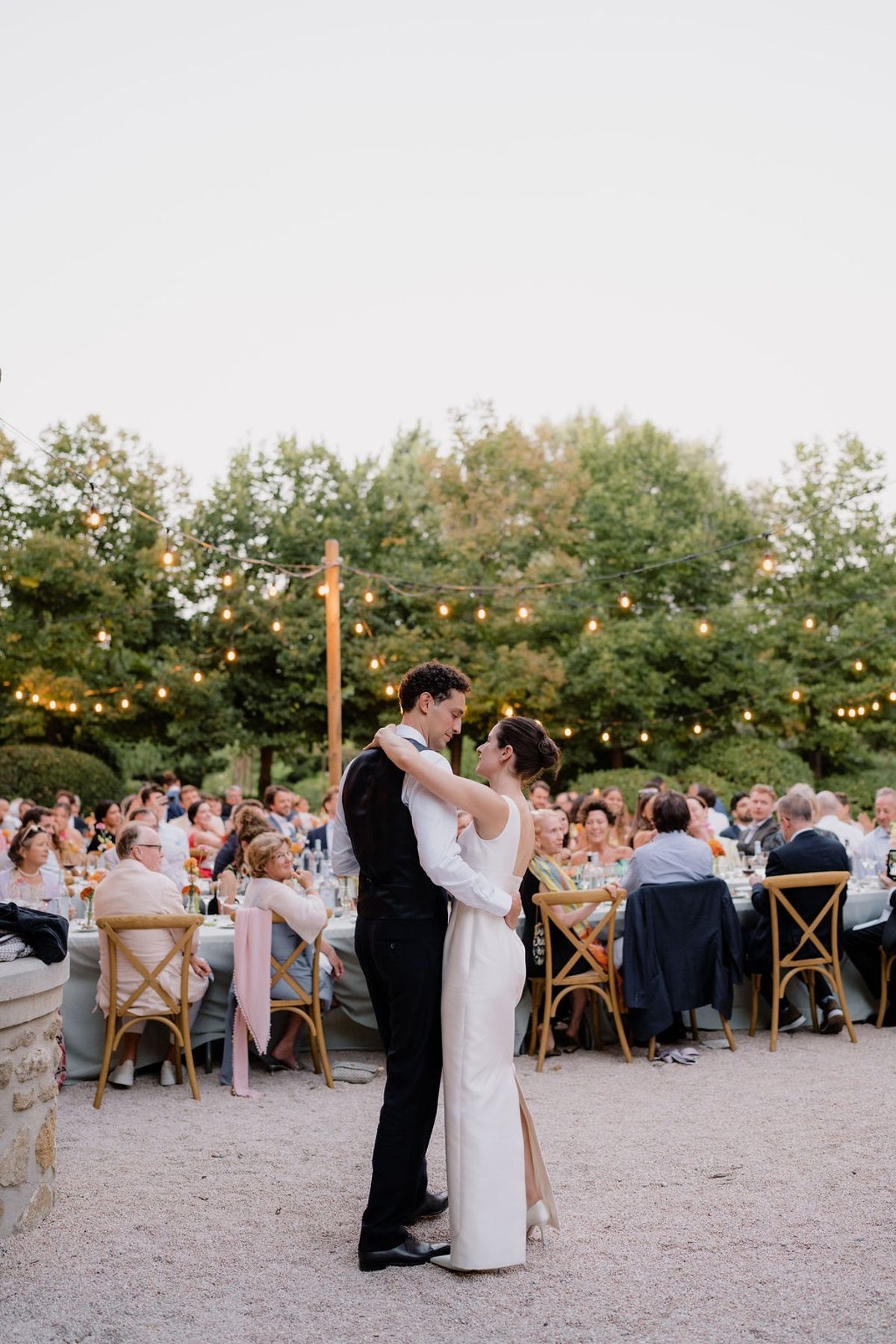 Newlyweds slow dancing on gravel terrace at twilight under bistro string lights