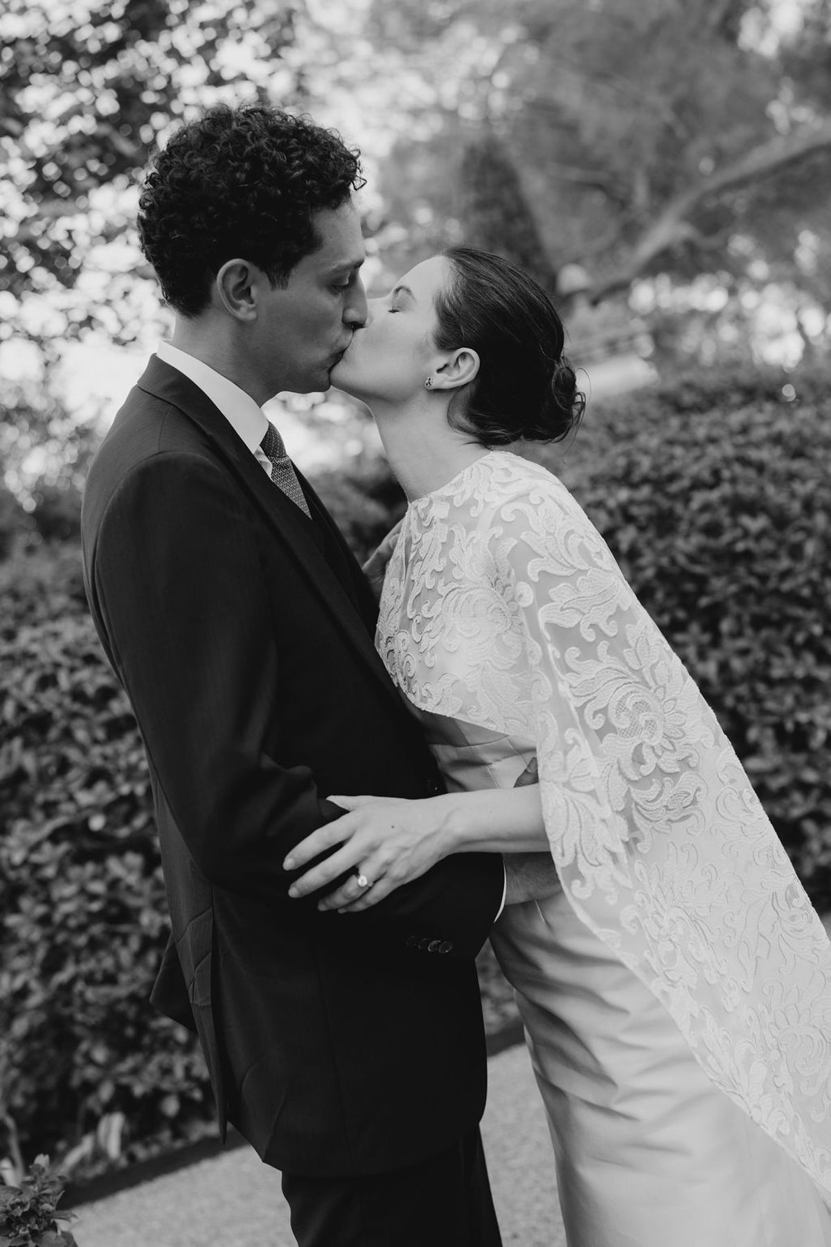 Close-up couple kiss in front of clipped hedge ring catching the light