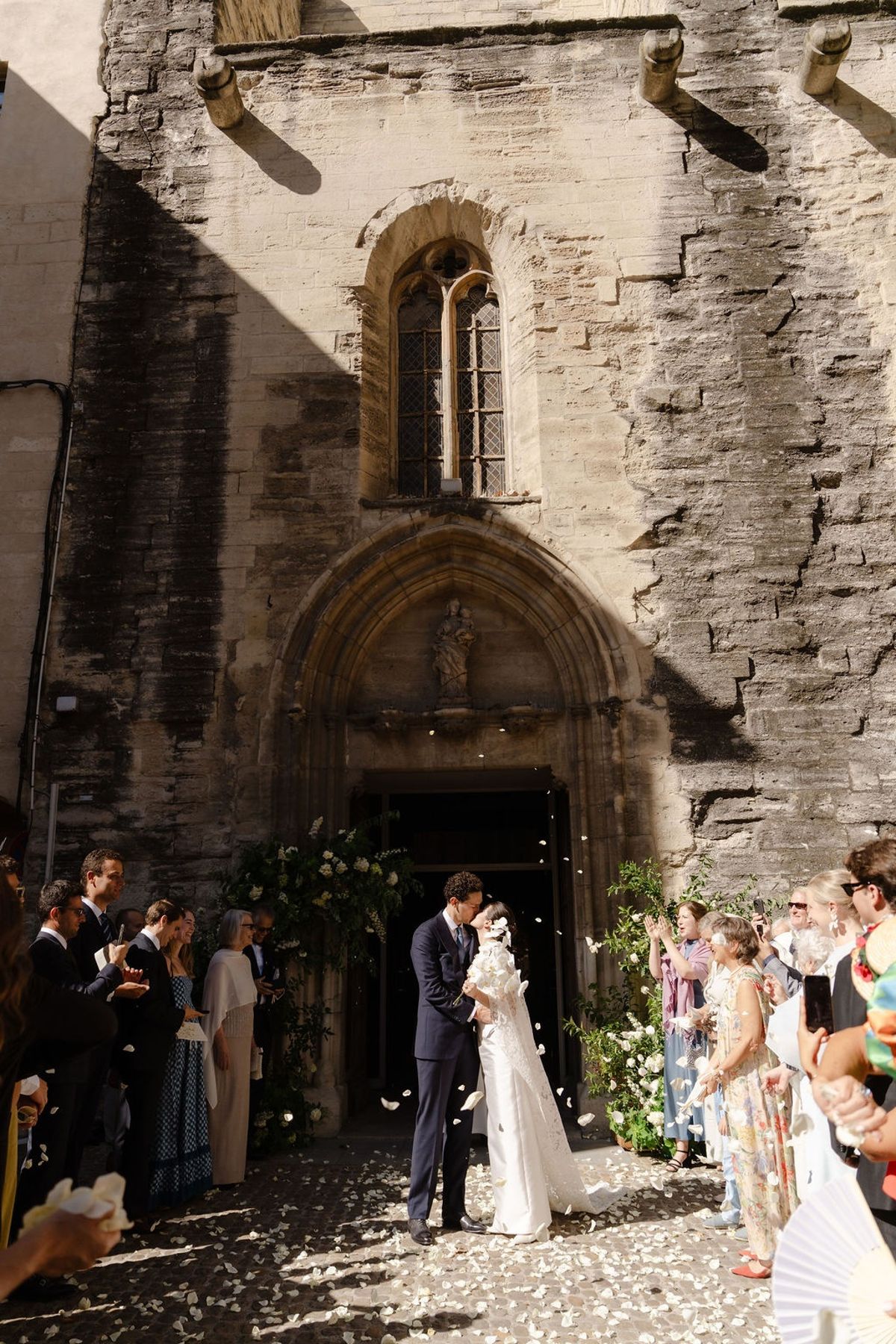 Newlyweds kiss in church doorway under petal toss from guests beside gothic arches