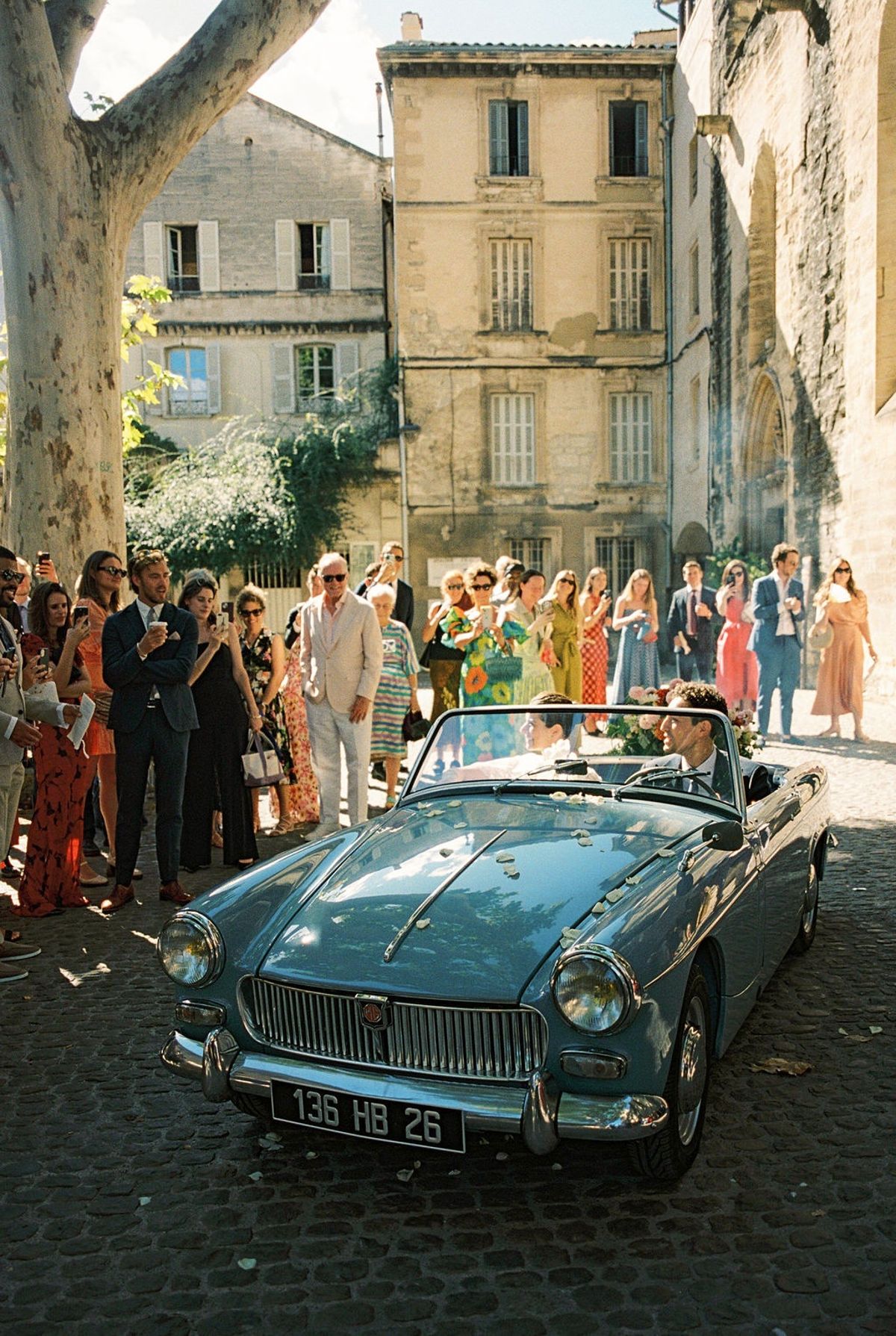 Couple seated in powder-blue convertible with plaza crowd watching under plane tree