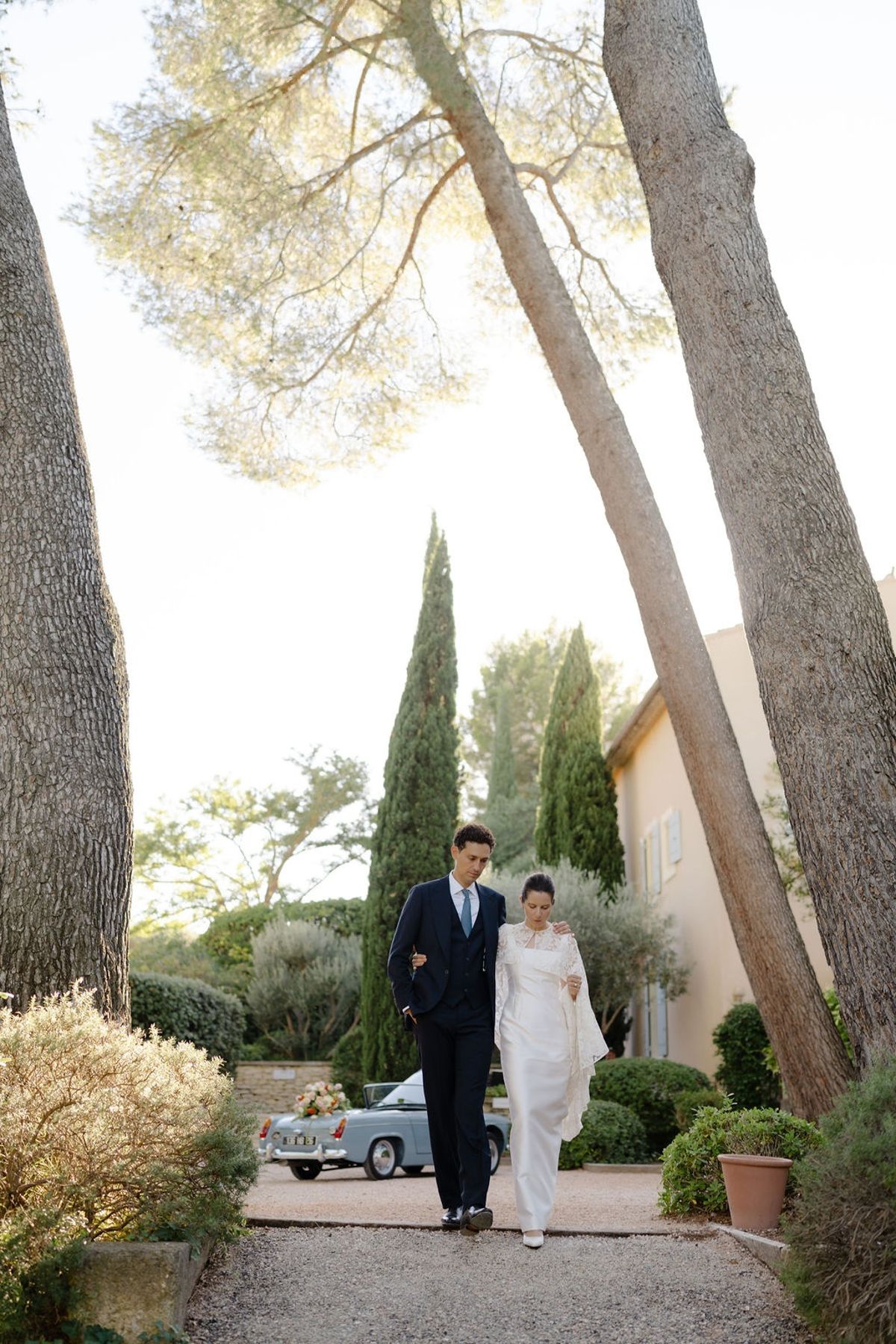 Couple walking arm in arm down gravel drive with parasol pines and convertible behind