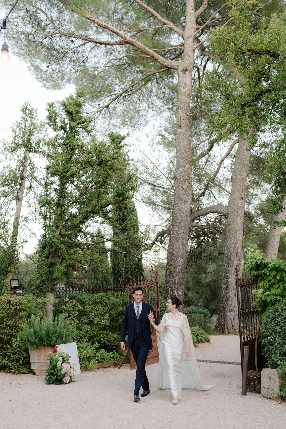 Couple walking through iron-gated garden entrance flanked by topiary and pines