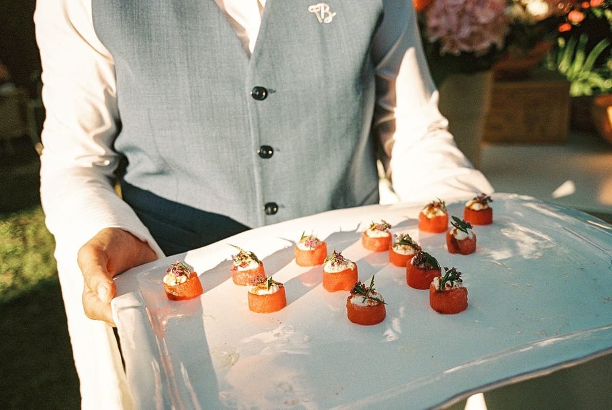 Waiter in grey waistcoat holding tray of watermelon canapes with cream and blossoms