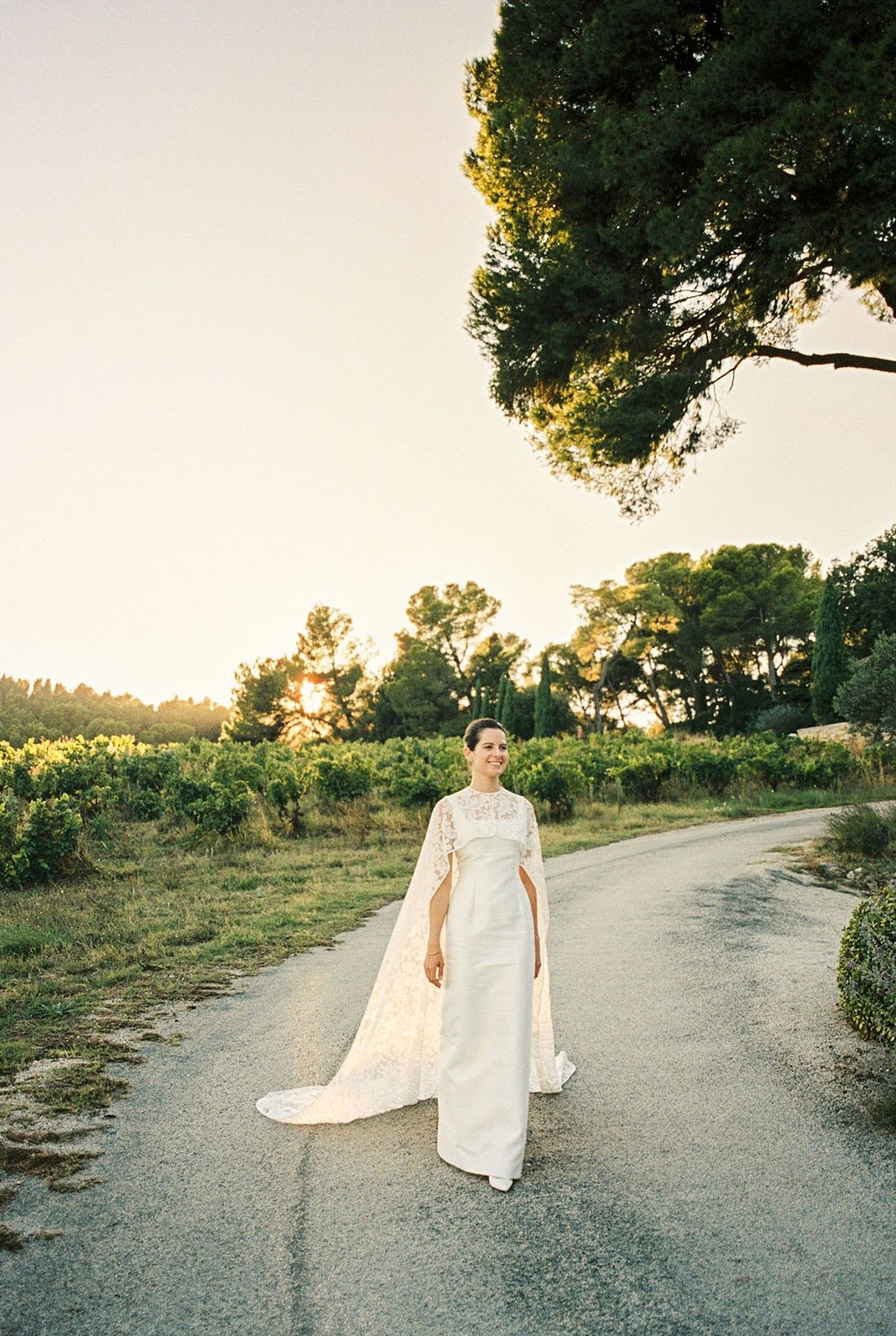 Bride walking alone down vineyard road in lace cape with golden sun on horizon