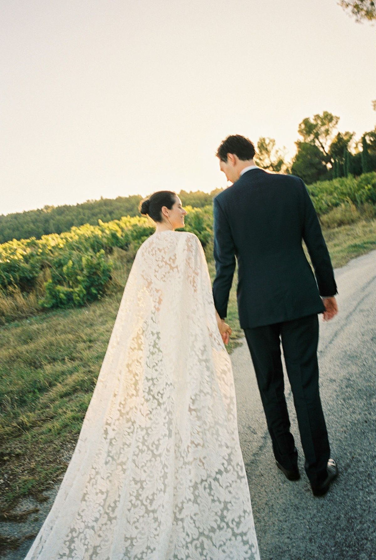 Couple from behind walking hand in hand down vineyard road with lace cape trailing