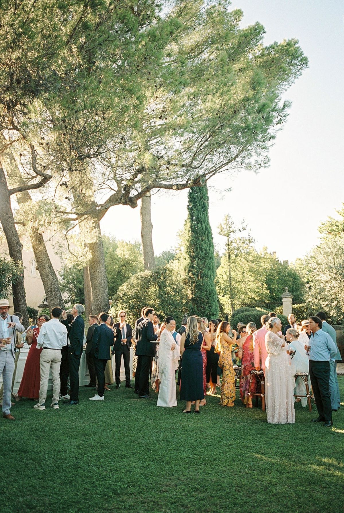 Wide view of cocktail hour on chateau lawn with newlyweds centre crowd in golden light