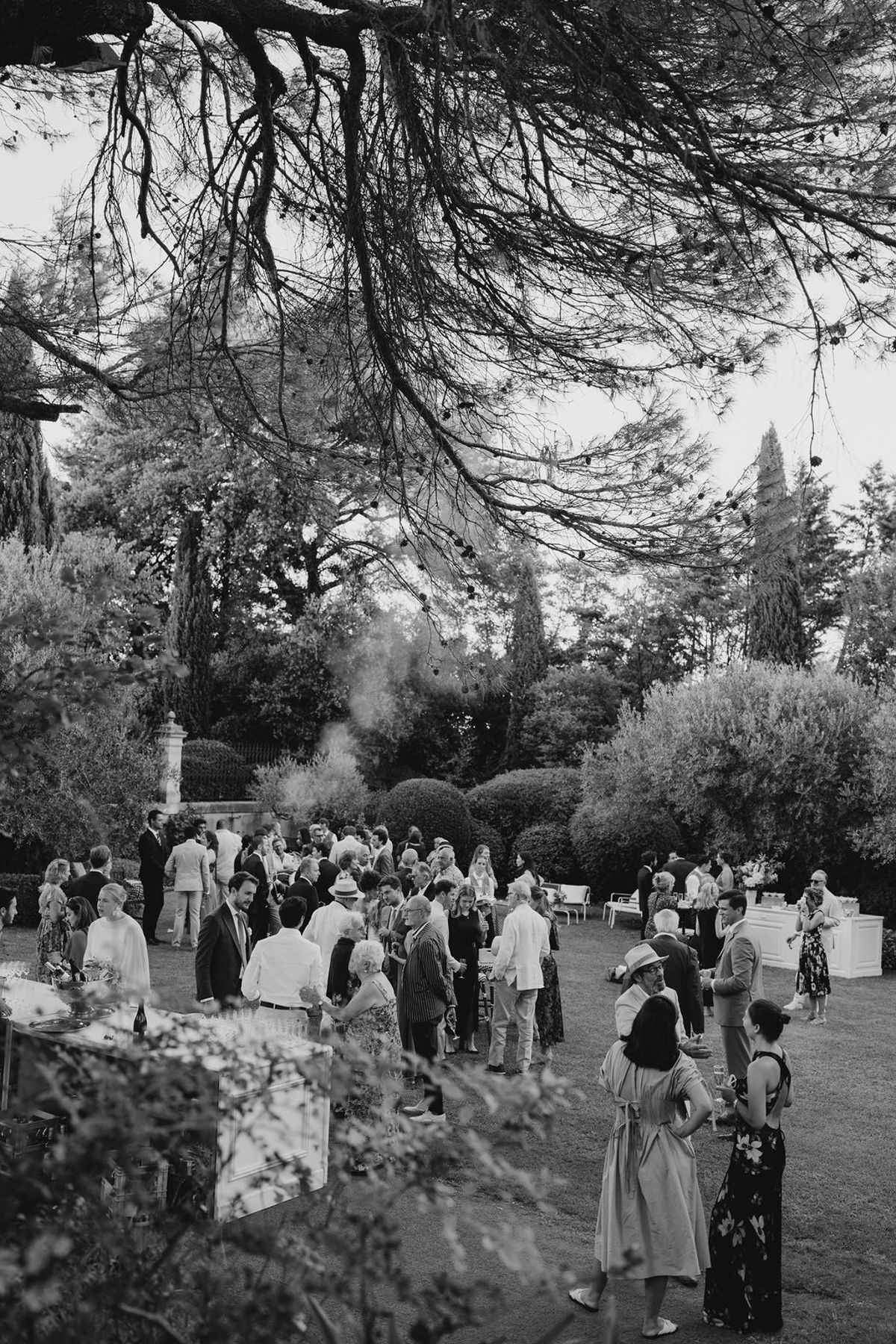 Black and white wide view of guests on chateau lawn under cedar canopy with drinks stations