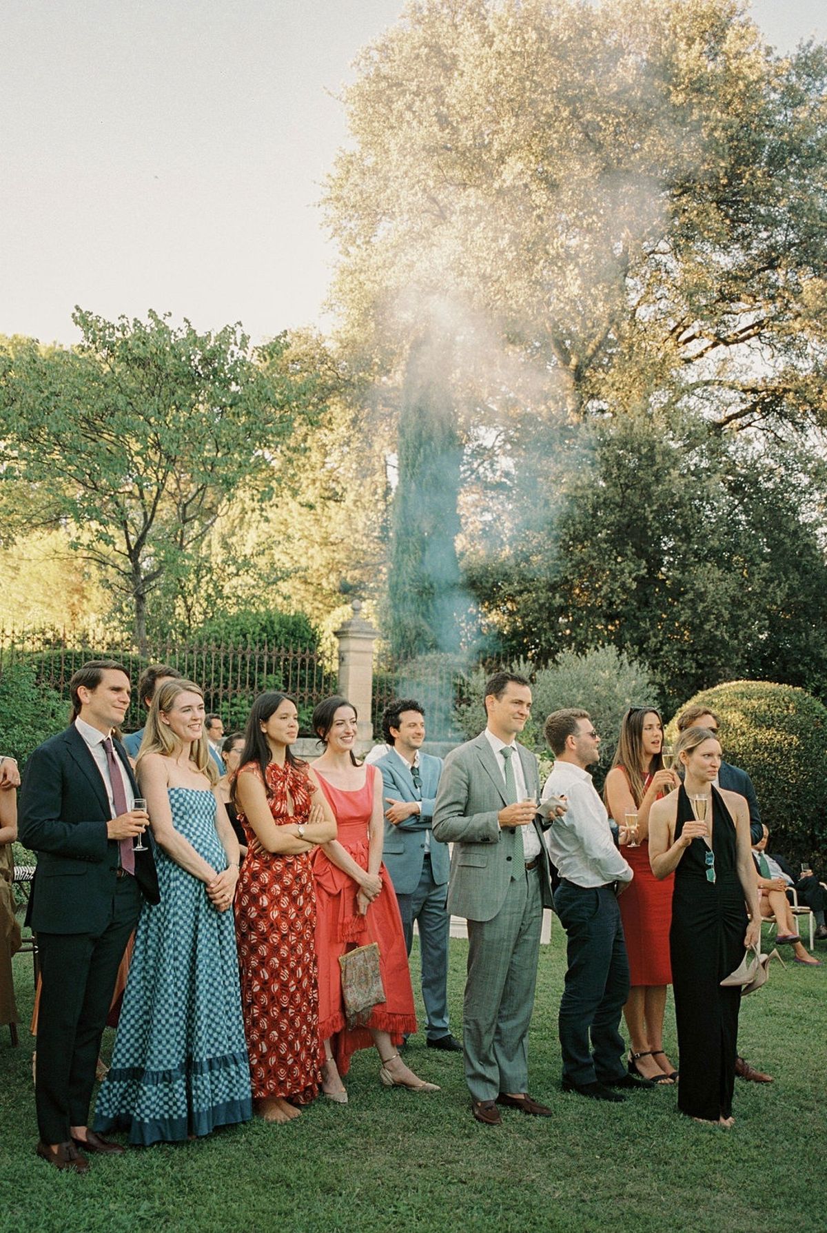 Guests in colourful vintage-inspired attire on lawn with drinks and sunlit trees behind