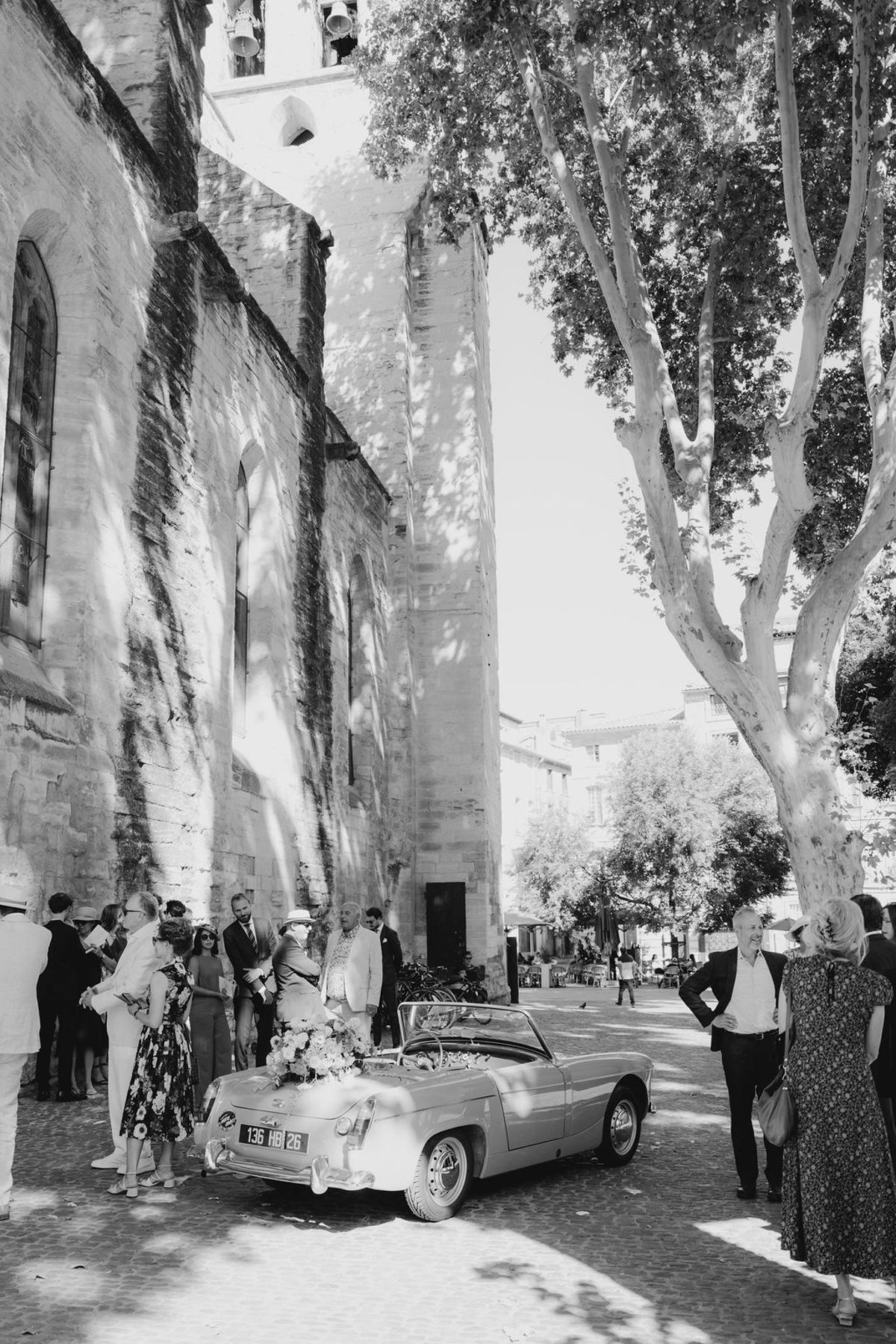 Black and white vintage convertible outside medieval church with guests under plane tree