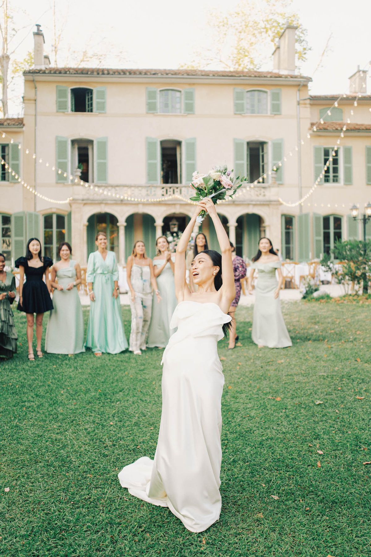 Bride tossing blush bouquet to eight female guests on lawn before ochre Provencal manor with fairy lights
