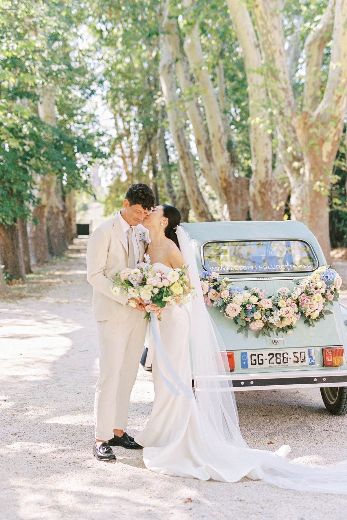 Couple kissing on tree-lined avenue bride in ivory gown with veil groom in sand suit mint-green 2CV behind