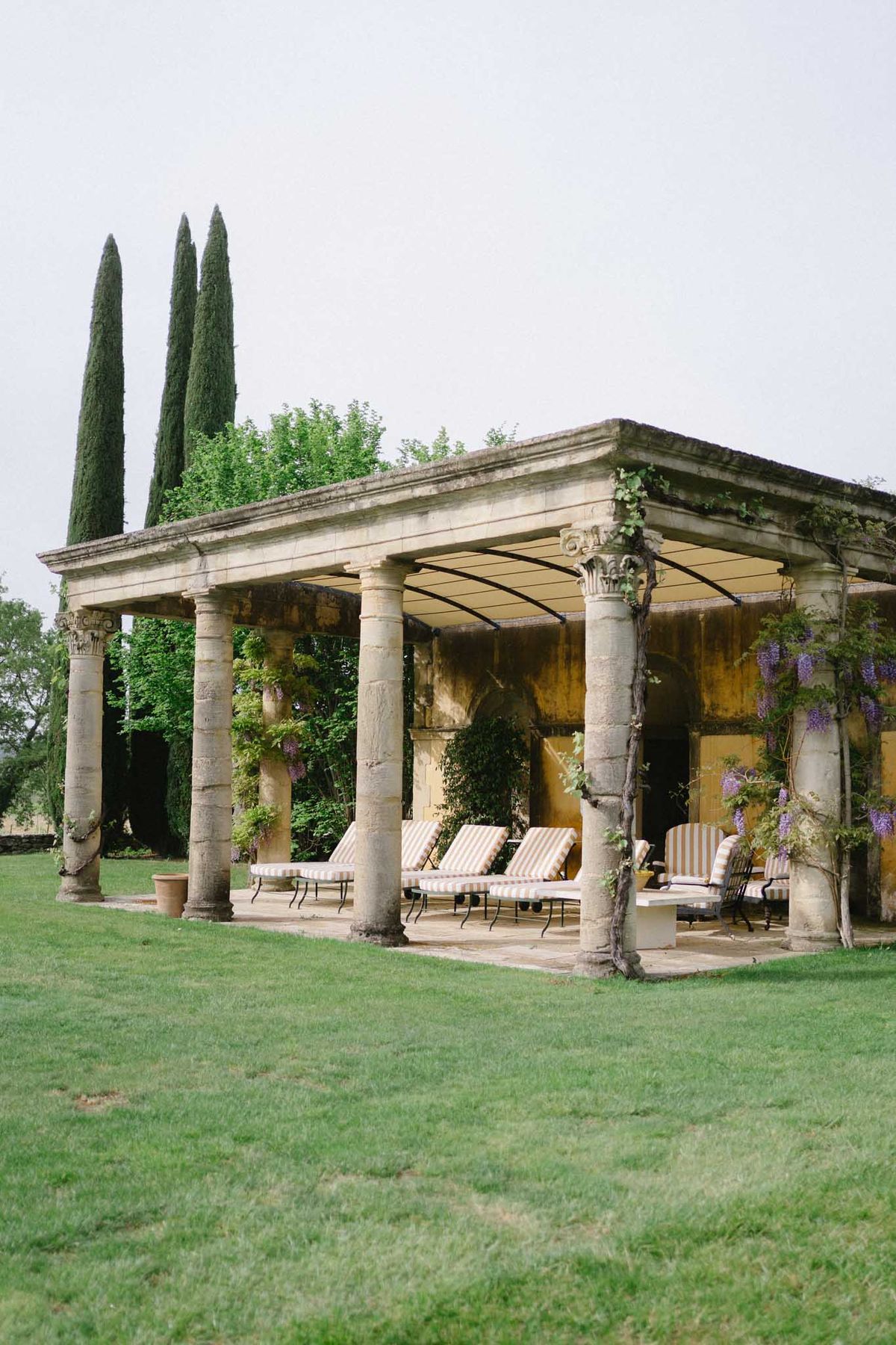 Stone Corinthian colonnade with canvas canopy, striped loungers, and purple wisteria climbing columns