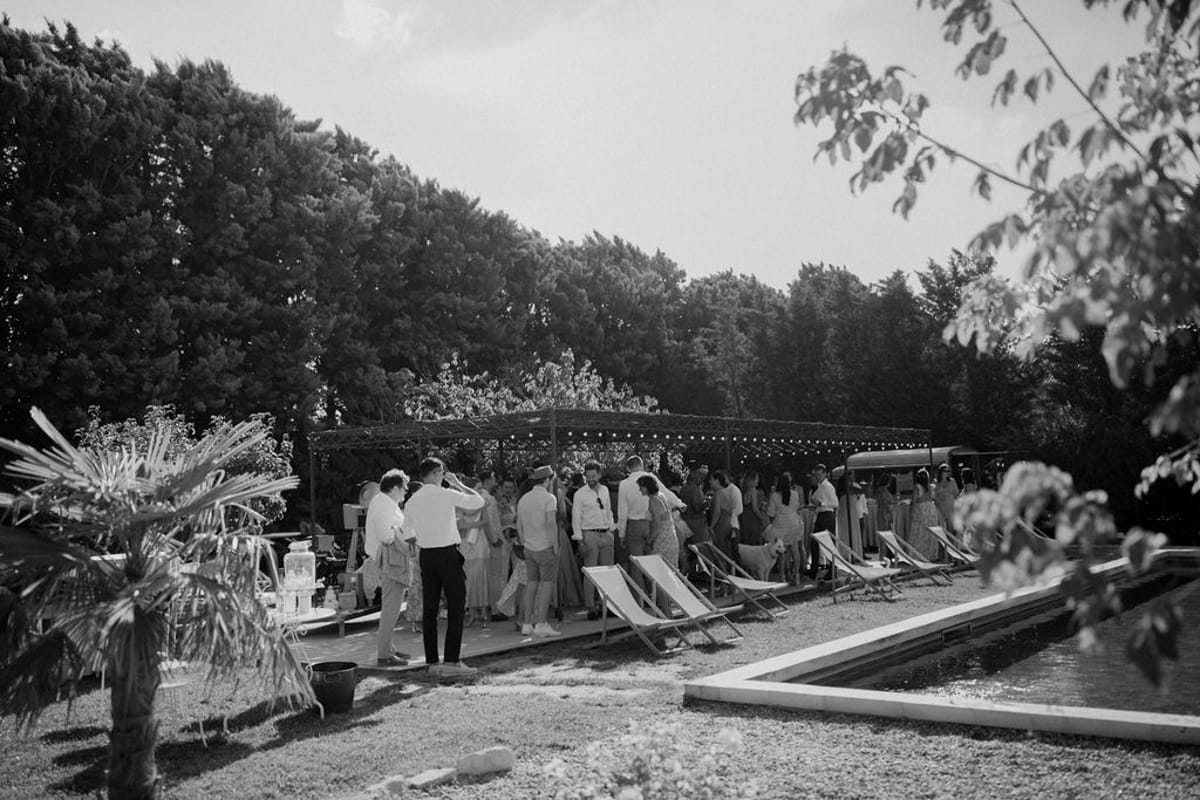 Black and white of guests mingling on poolside wooden deck with pergola and globe fairy lights in daylight