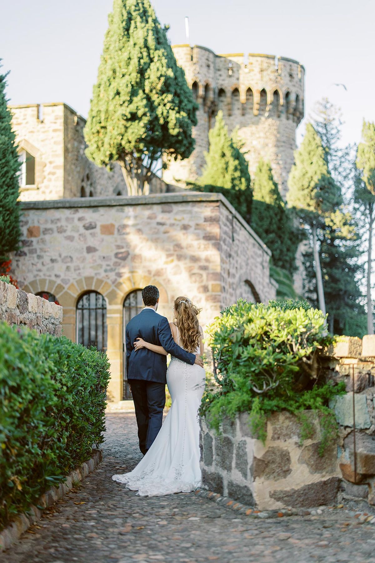 Bride and groom walking away along cobblestone path toward medieval castle with crenellated tower in late afternoon light