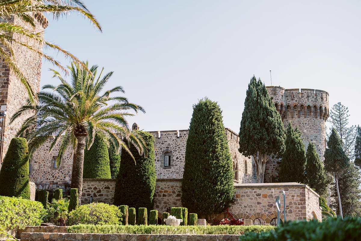Stone castle wedding venue with rounded tower, battlements, topiary hedges, cypress trees, and palm tree in bright daylight