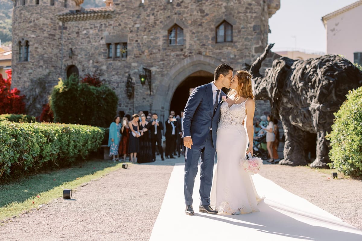 Couple kissing on white aisle before medieval stone castle with guests watching in background